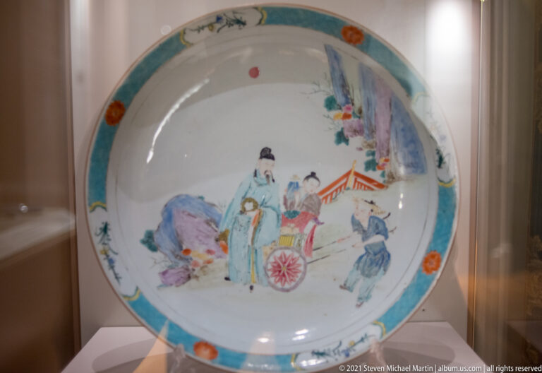 Shallow porcelain dish with turquoise rim and a multicolor Chinese garden scene of robed figures, rocks, plants, and a pavilion.