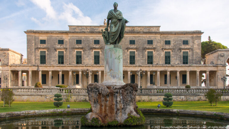 Wide garden view of the palace façade behind a seated bronze monument, clipped shrubs, stone balustrade, and reflecting pool.