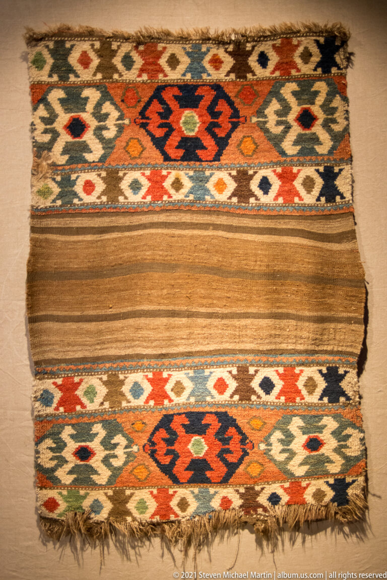 Rectangular woven textile with a plain brown center, colorful geometric border bands, angular medallions, and short fringe.