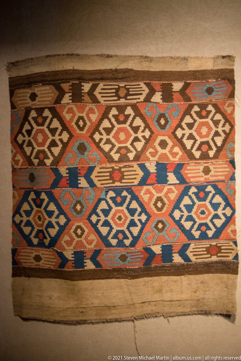 Rectangular woven textile with rows of multicolored hooked diamonds and angular geometric motifs, displayed flat behind glass.