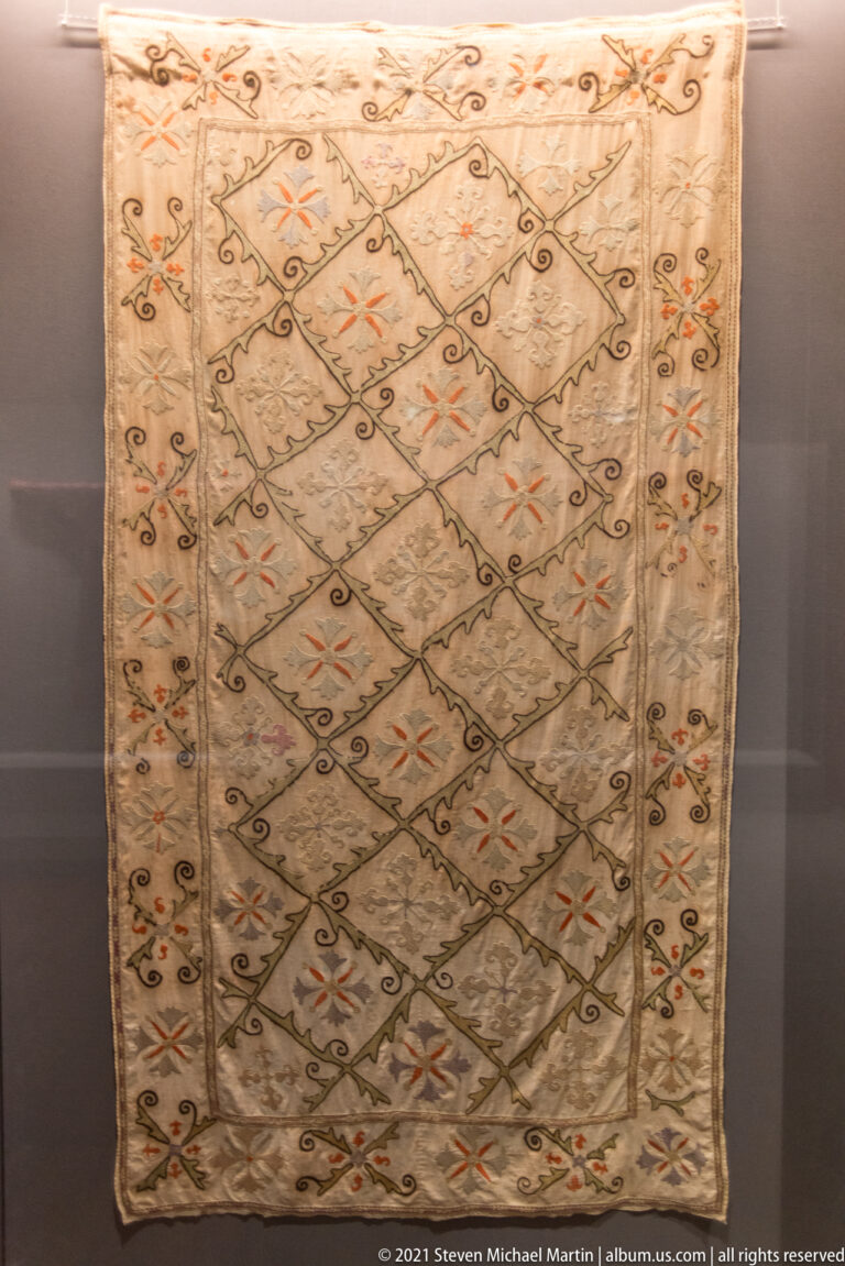 Pale embroidered textile with diagonal lattice, small floral motifs, orange accents, and a narrow patterned border.