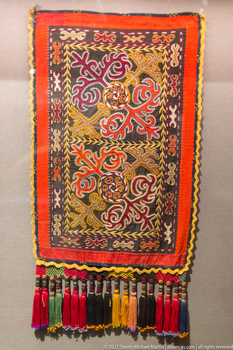 Red decorative textile with curving multicolored motifs, narrow patterned borders, and a row of long tassels.