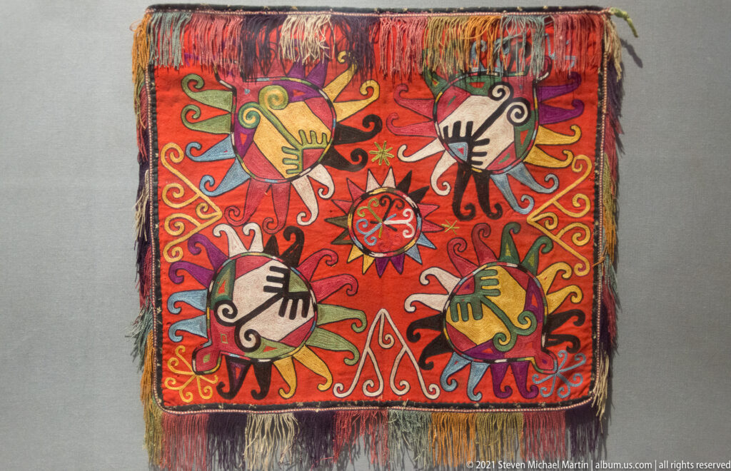 Square red textile with four stylized human figures around a central rosette, bordered by multicolored fringe.