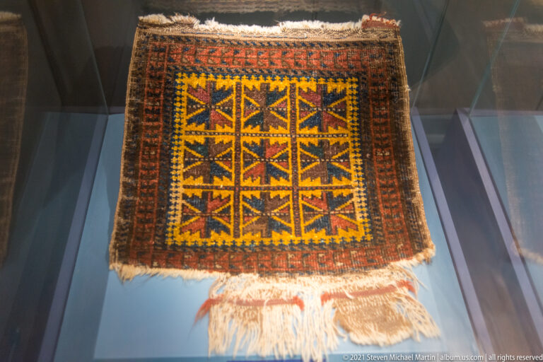 Small fringed rug with a yellow center containing nine angular geometric motifs and dark rust-brown borders.