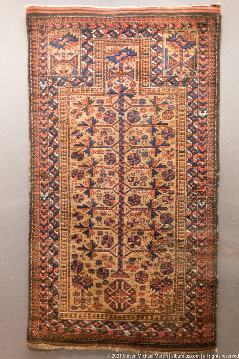 Rectangular patterned rug with a stepped niche-like upper field, dense small motifs, and multiple rust and blue borders.