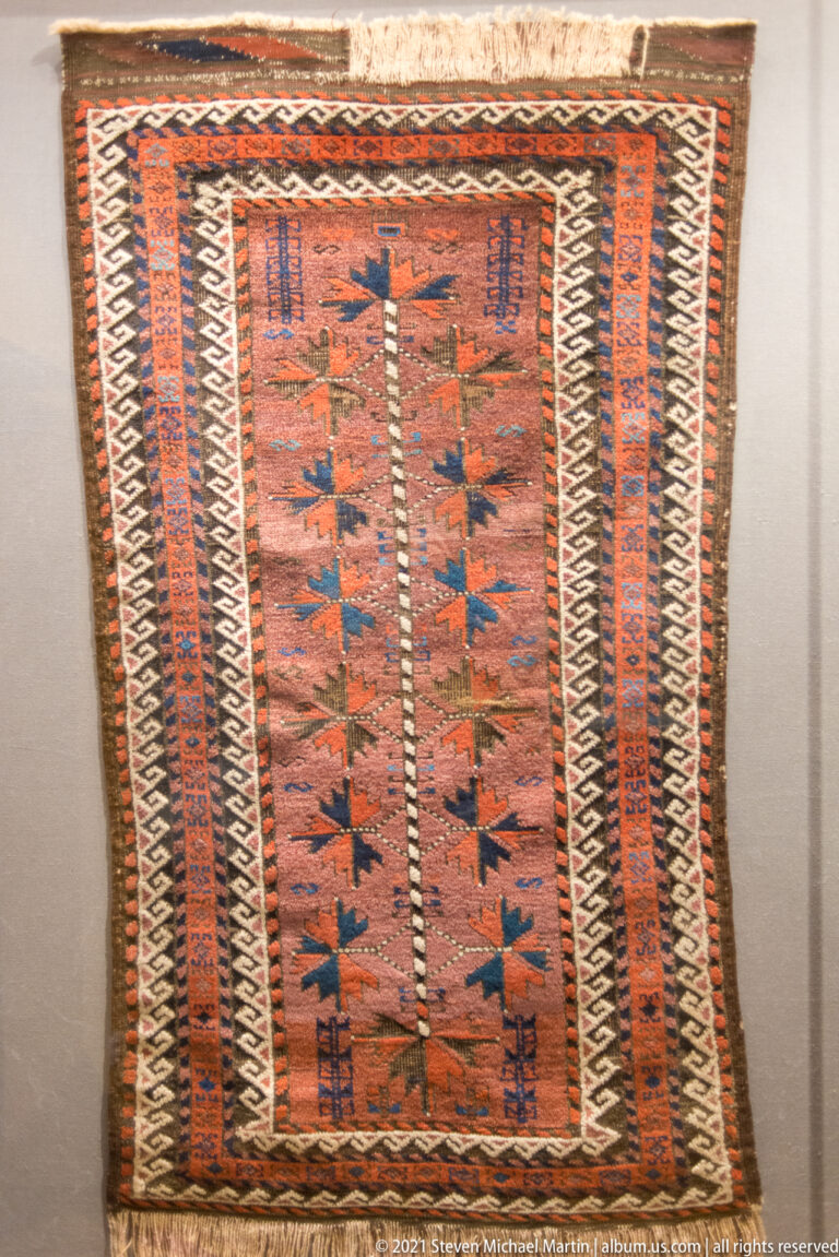 Long narrow rust-colored rug with repeated dark angular motifs and parallel striped and zigzag borders.