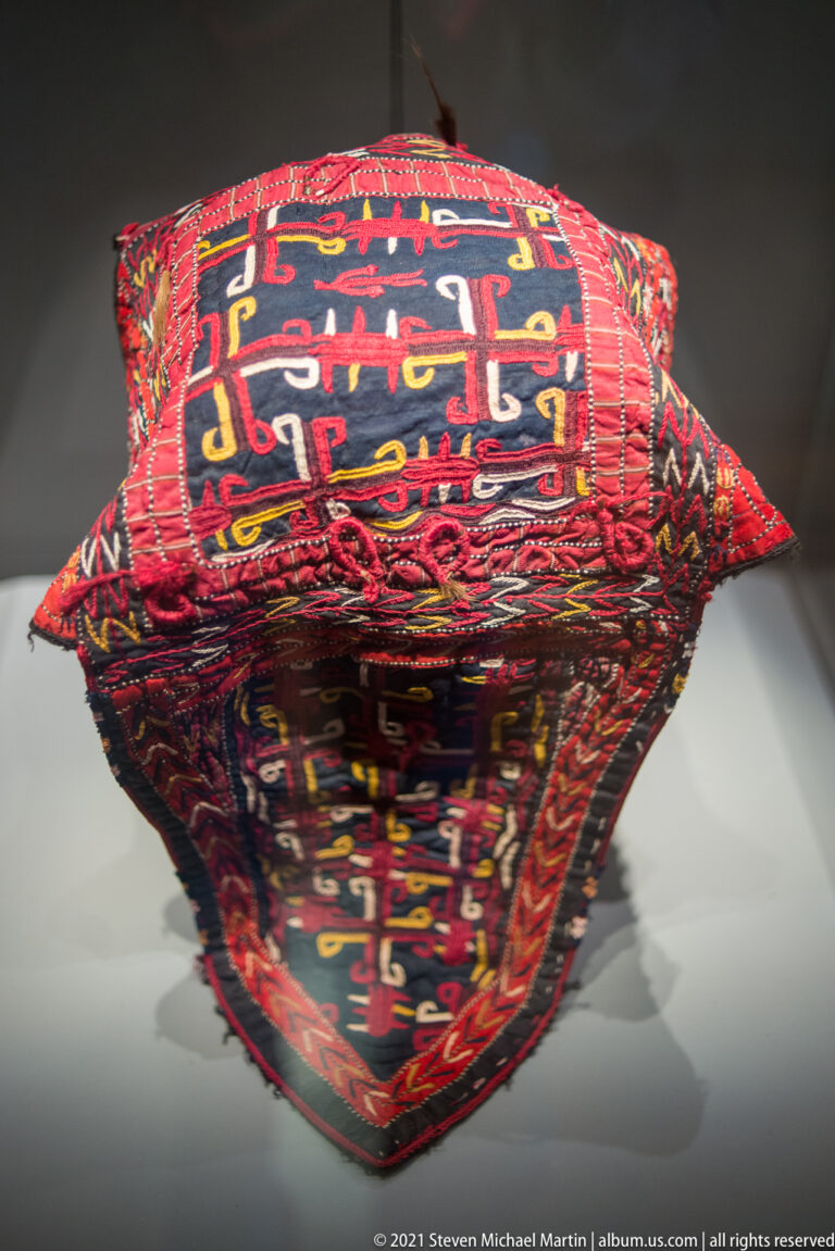 Red padded textile object with dense multicolored curvilinear patterns and pointed ends, reflected on a glass surface.