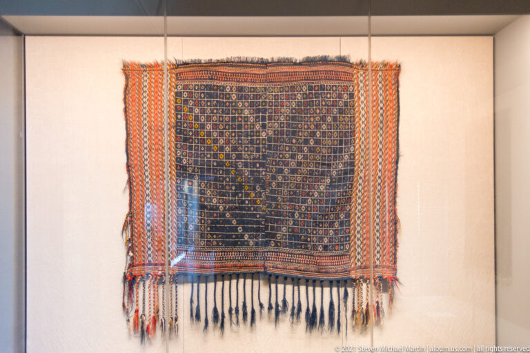 Large dark woven hanging with orange and blue patterned side bands and a row of long dark tassels.