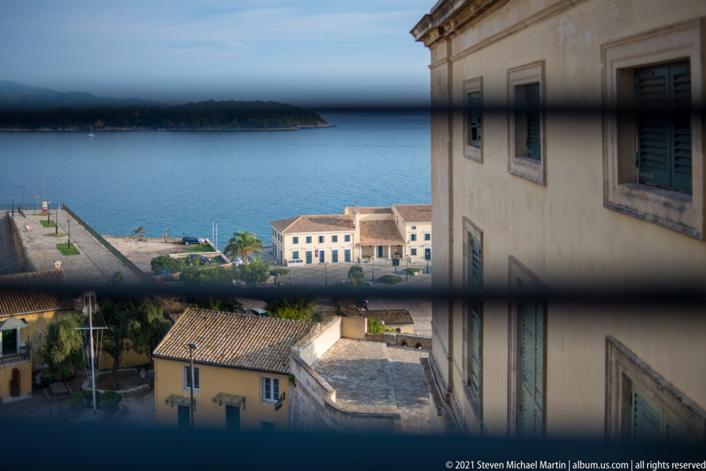 Corfu waterfront and blue sea seen through window bars beside the pale stone exterior of the museum building.