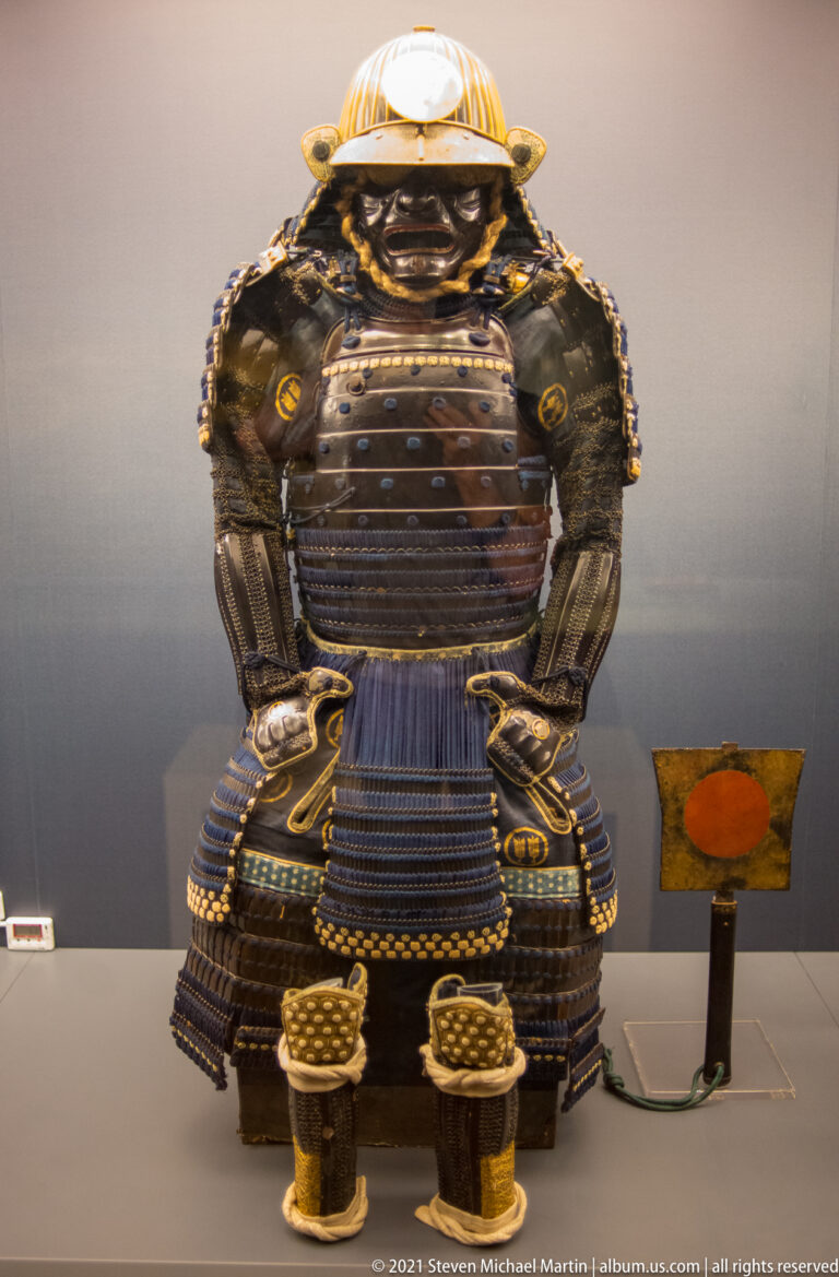 Front-facing Japanese samurai armor with helmet, dark lamellar plates, blue lacing, gold fittings, and armored footwear.