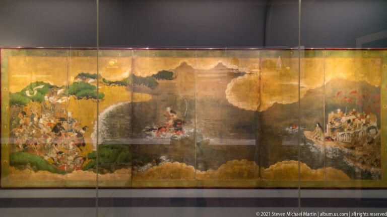 Large Japanese folding screen on a gold ground with groups of figures, buildings, trees, and cloud forms across several panels.