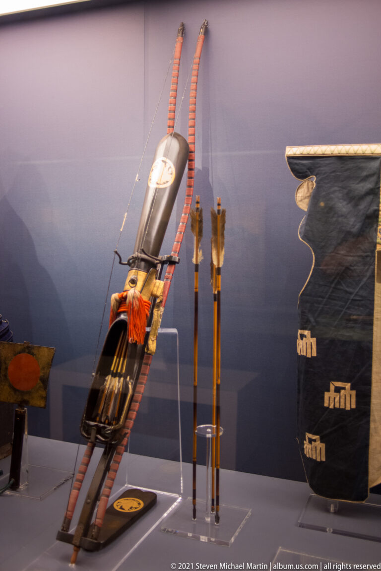 Japanese archery equipment including a tall bow, dark quiver with red bindings, and several arrows displayed upright.