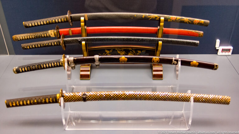 Japanese swords and scabbards displayed horizontally, with black and red lacquer, patterned grips, guards, and metal fittings.