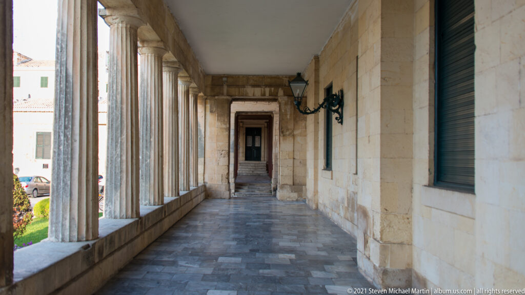 Long palace colonnade with stone columns, gray paving, green shutters, wall lamps, and a doorway at the far end.