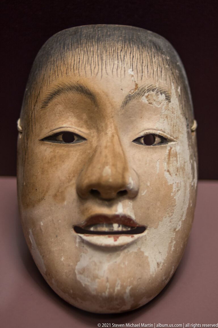 Pale Japanese theatrical mask with narrow eyes, small parted lips, painted hairline, and areas of worn surface.