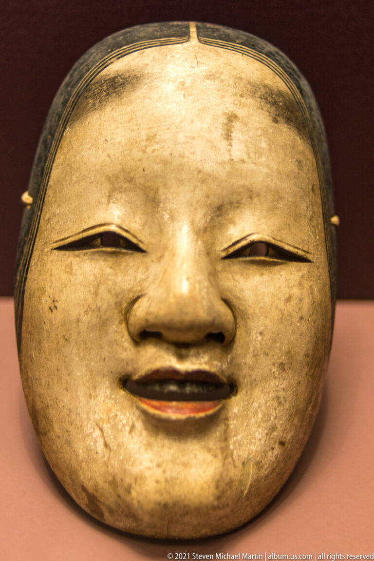 Rounded gold-toned Japanese theatrical mask with narrow eyes, softly modeled cheeks, and a small smiling mouth.