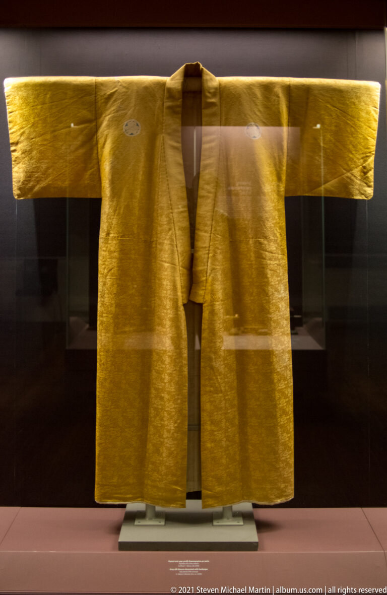 Gold-colored Japanese robe with broad sleeves, subtle woven decoration, and small circular crests near the shoulders.