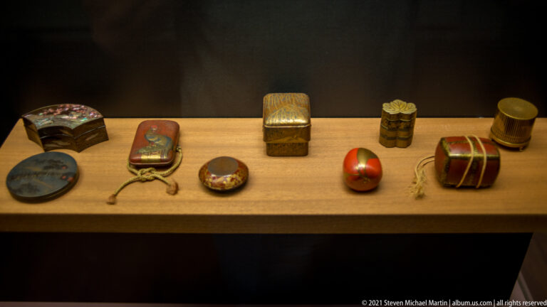 Assorted small Japanese lacquer boxes and containers in red, black, and gold, arranged across a museum shelf.