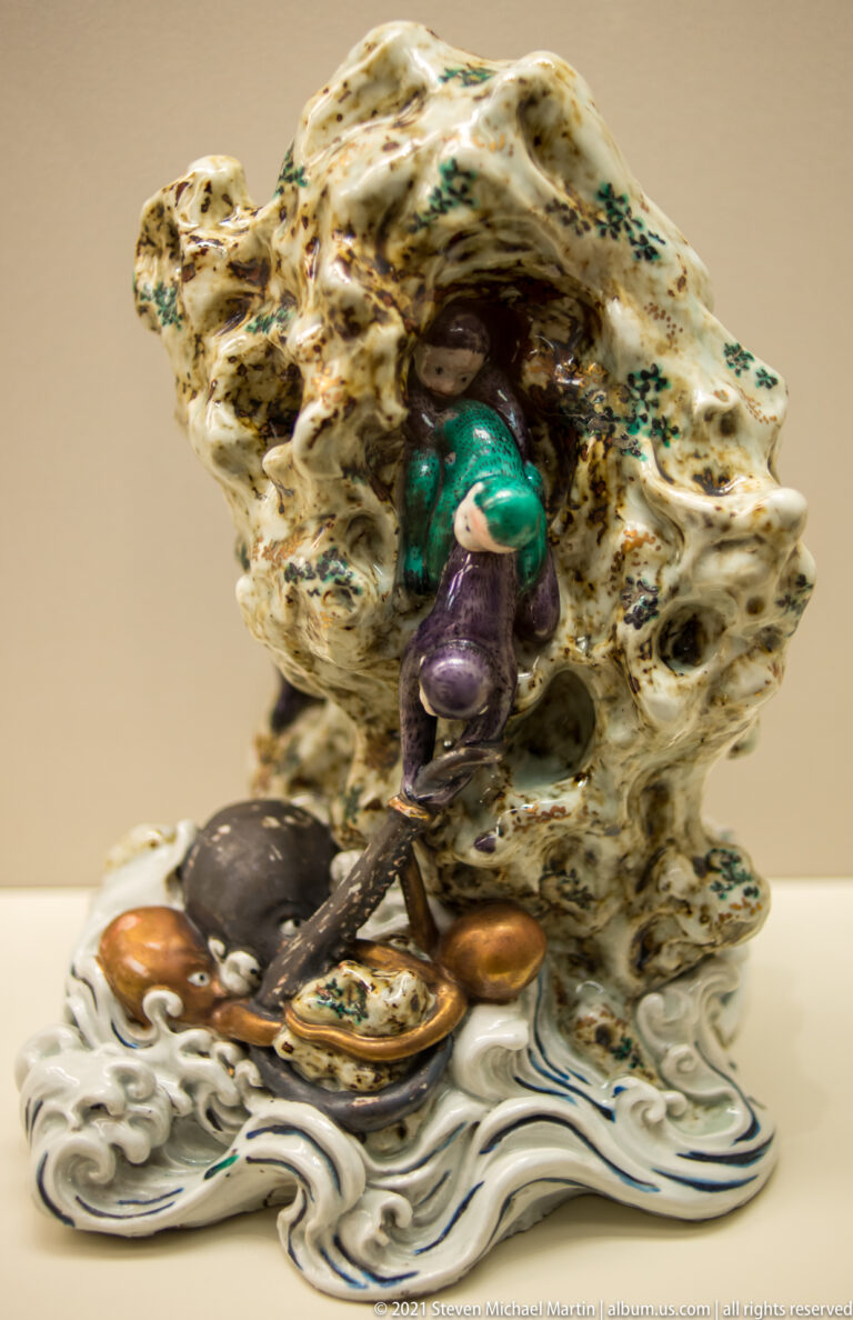 Glazed ceramic group with a small colorful figure beside a pierced rock formation rising from stylized waves.