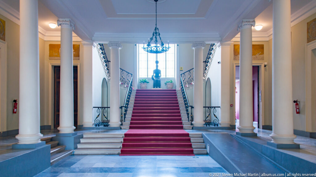Symmetrical palace hall with white columns, gray stone floor, red-carpeted central staircase, iron railings, and hanging lantern.