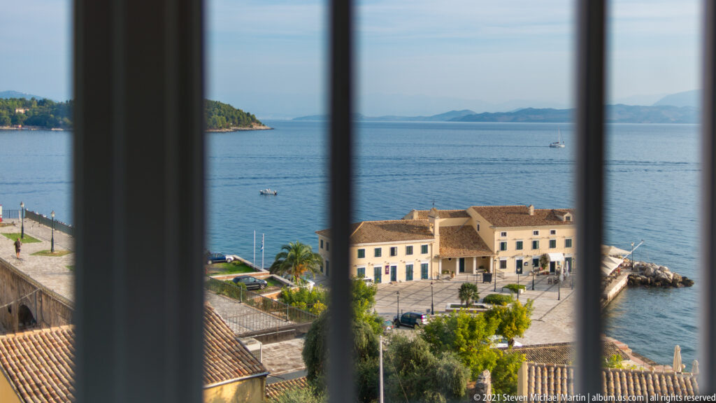 Corfu waterfront, pale seaside building, terracotta roofs, and blue Ionian Sea seen through vertical window bars.