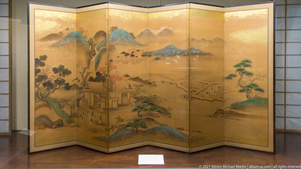 Gold-ground folding screen with mountains, pine trees, a small settlement, and winding watercourse across multiple panels.