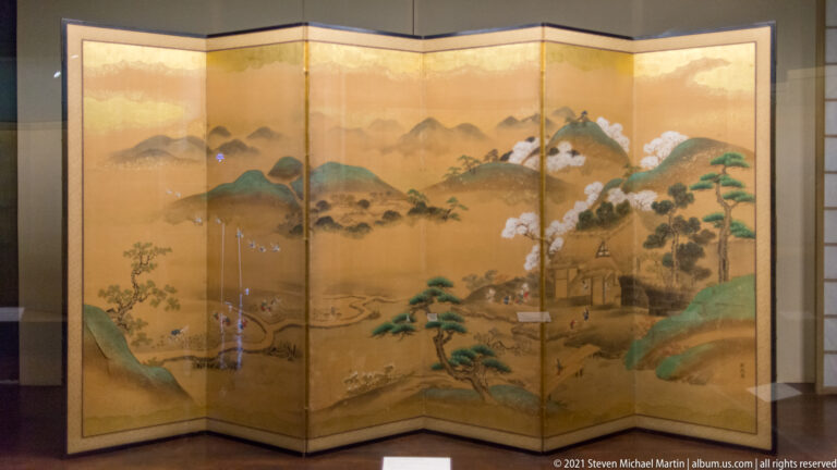 Gold folding screen with distant mountains, pine trees, white cloud forms, and buildings concentrated toward the right.