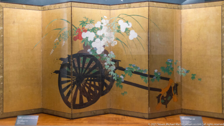 Gold-ground screen with a dark wooden cart carrying white and red blossoms, green foliage, and long projecting shafts.
