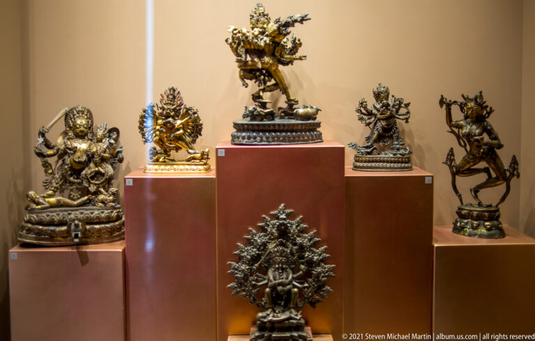 Stepped display of small bronze devotional sculptures, including seated and dancing figures with crowns, multiple arms, and halos.