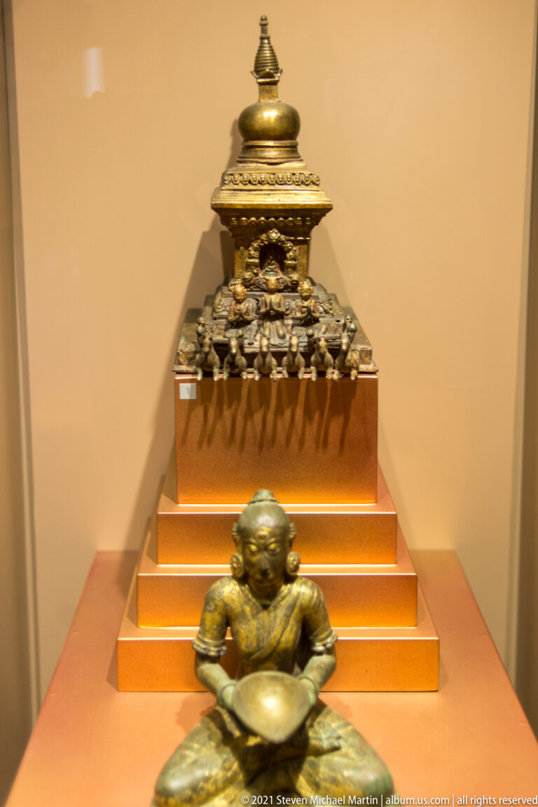 Seated bronze figure in front of a tiered metal shrine or reliquary topped by a finial and ornate projecting details.