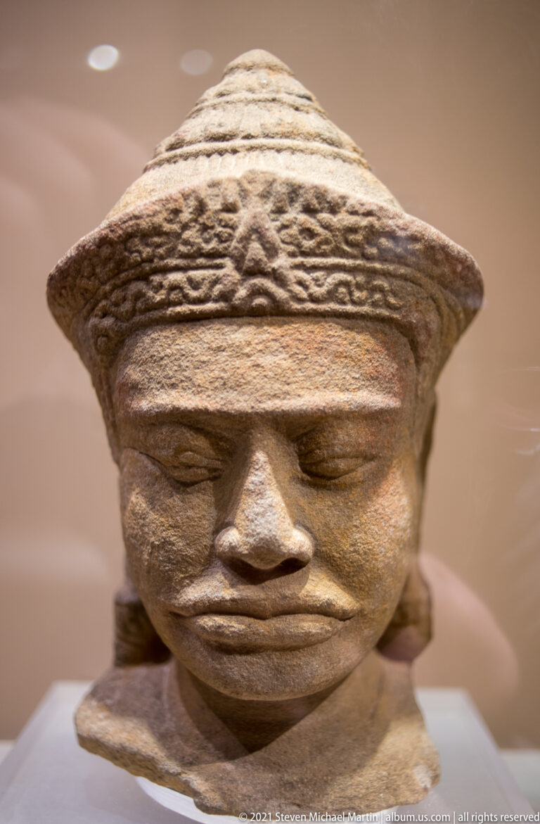 Stone Cambodian Buddha head from the Angkor period with broad face, downcast eyes, full lips, and patterned headdress.