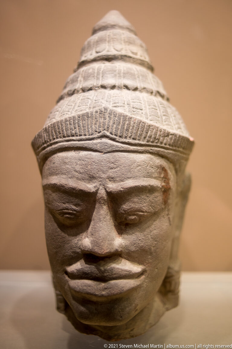 Stone Angkor-period Cambodian Buddha head with heavy-lidded eyes, broad nose, slight smile, and tall tiered headdress.