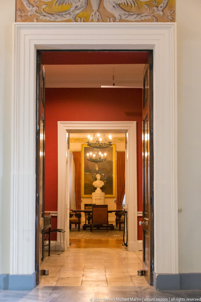 Open double doors reveal aligned palace rooms with red walls, marble floor, draperies, ceiling decoration, and a distant white bust.