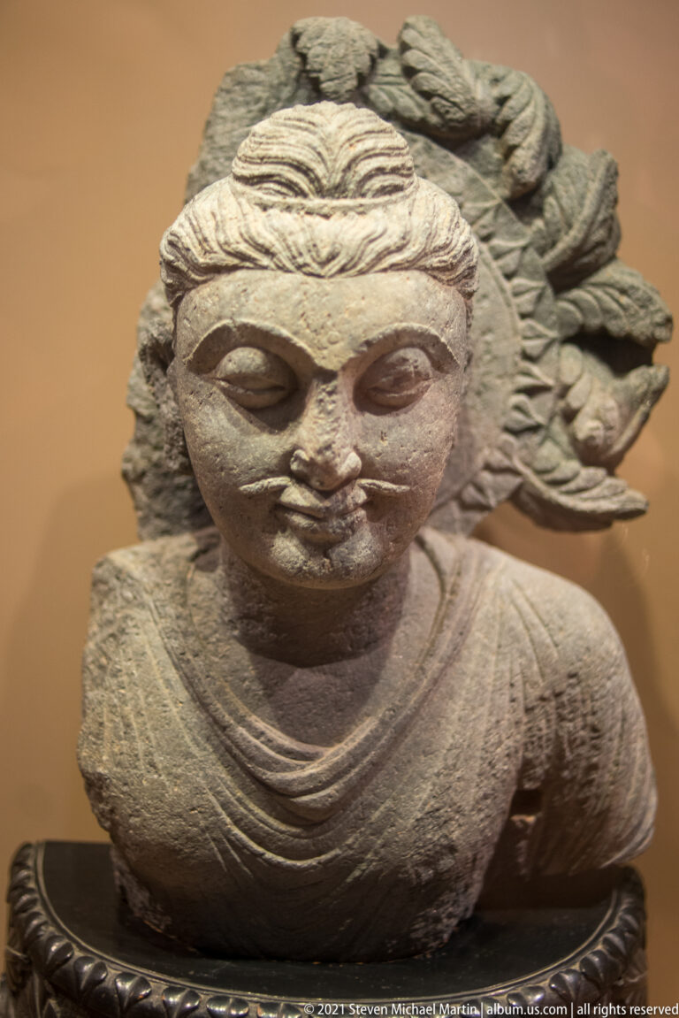 Gandharan grey-schist bust of Hariti with wavy hair, floral headdress, necklace, draped garment, and softly modeled face.