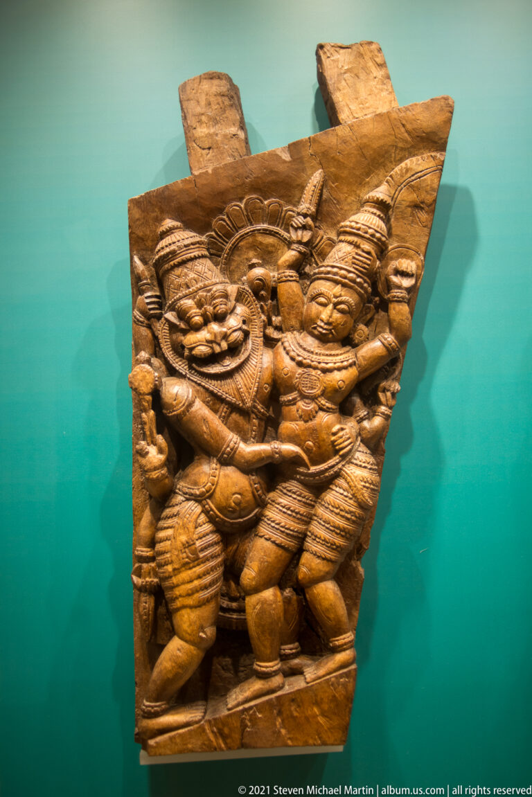 Wooden relief with two elaborately dressed standing figures wearing tall headdresses and jewelry within a narrow frame.
