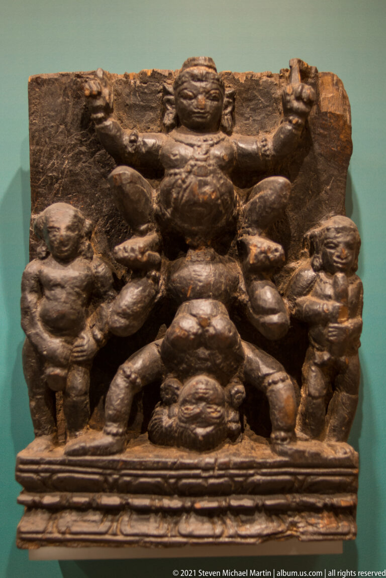 Horizontal carved wooden relief with a large central figure surrounded by several smaller human figures and an ornamental base.
