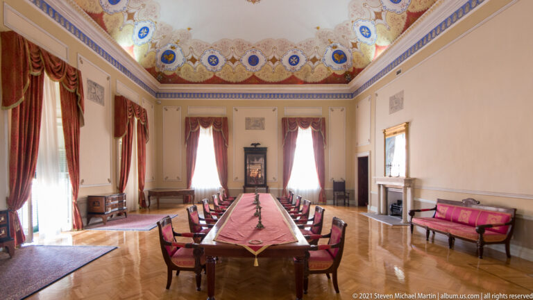 Long formal table with red chairs in an ornate palace room with tall draped windows, parquet floor, and decorated ceiling.