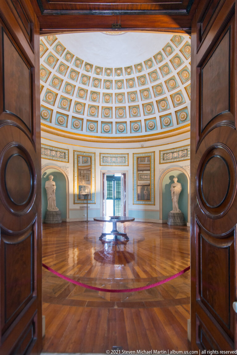 View through wooden doors into a circular palace rotunda with coffered dome, arched bust niches, and polished parquet floor.