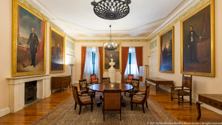 Ionian Senate Meetings Hall with original round table and chairs, large gilt-framed portraits, marble fireplace, and sculptural bust.