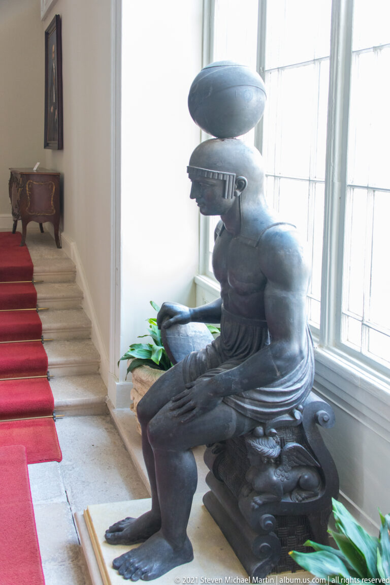 Dark seated male sculpture beside a red-carpeted staircase, leaning beneath a cushion-like form near tall bright windows.