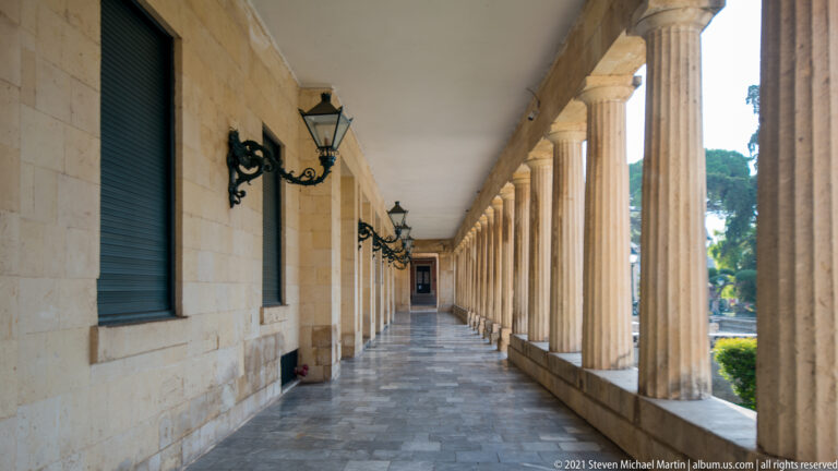 Long stone colonnade with repeated columns, wall lanterns, shuttered windows, and a tiled passage receding into the distance.