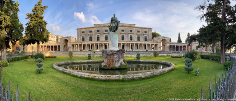 Palace of St. Michael and St. George behind a formal garden with circular basin and bronze statue on a stone pedestal.