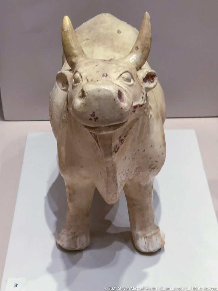 White-glazed ceramic bovine-shaped vessel with horns, four short legs, rounded muzzle, and an opening in its back.