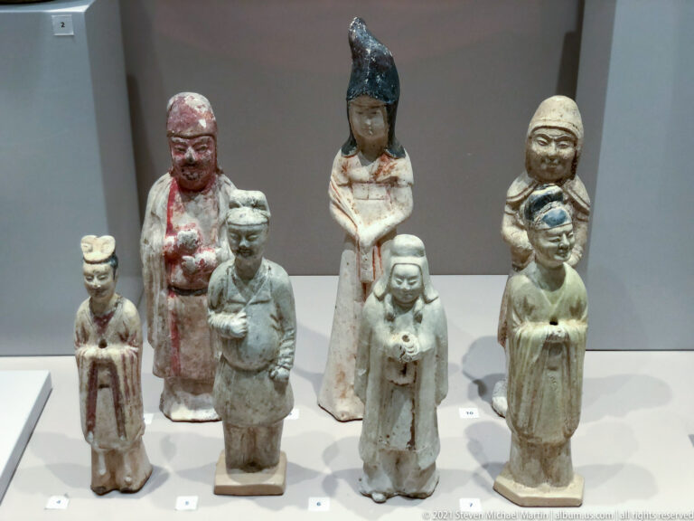 Six small painted ceramic figures in long robes, with varied hats, hairstyles, gestures, and surviving red, green, and cream pigment.