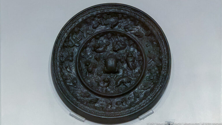 Circular dark bronze mirror with central boss and concentric bands of raised relief ornament on its decorated back.