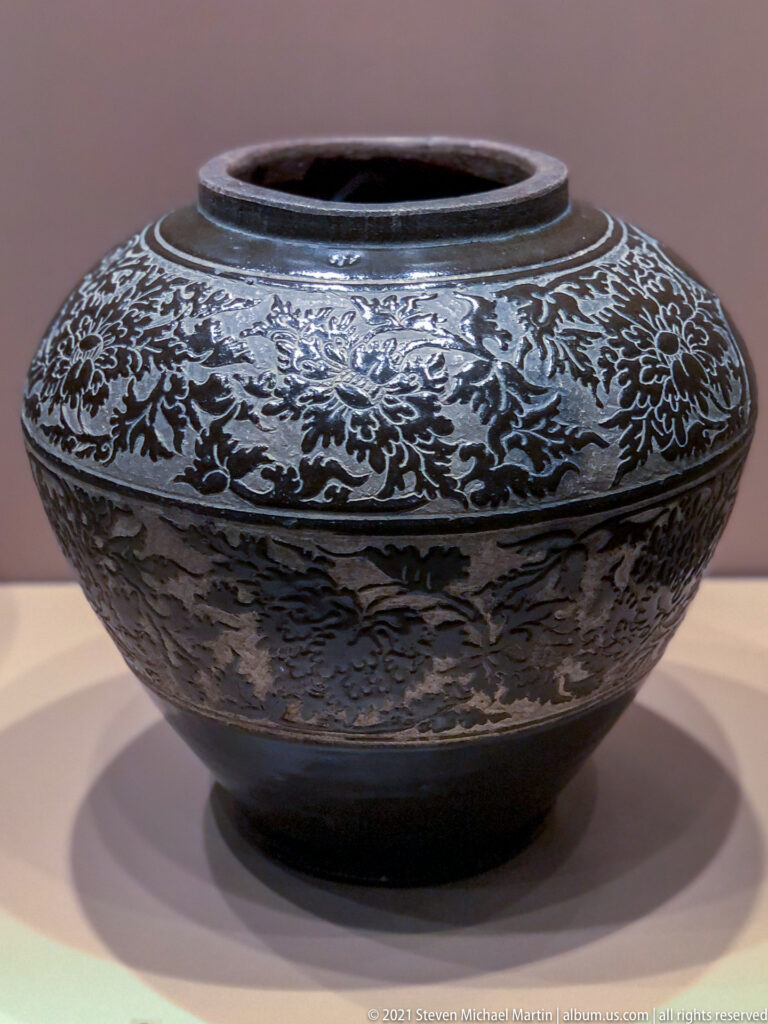 Wide low black-glazed vessel with open mouth, short foot, and dense pale floral scroll decoration around the body.