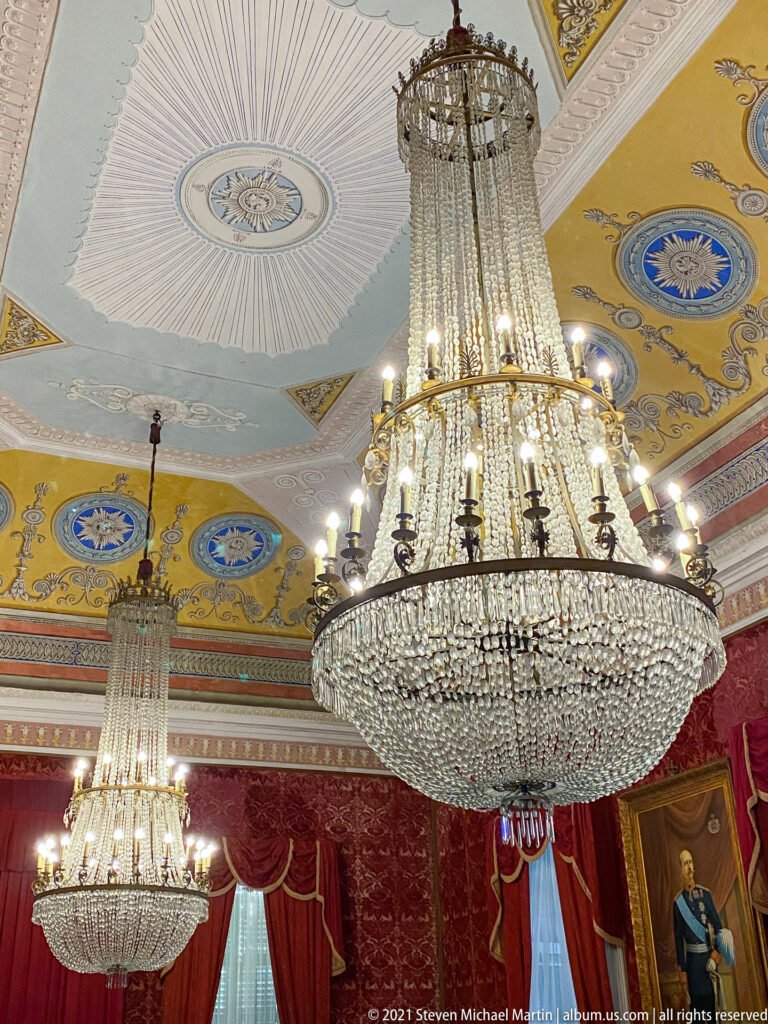 Two large crystal chandeliers hanging beneath the decorated Throne Room ceiling, with red draperies and portraits visible below.