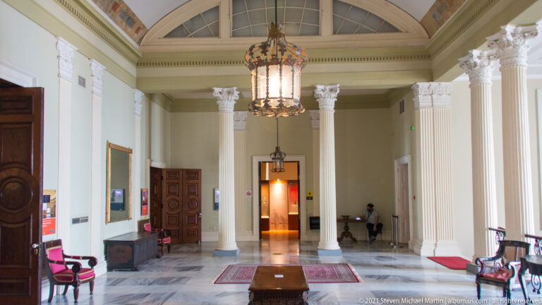 Tall museum hall with white columns, high glazed ceiling, hanging lantern, polished stone floor, wooden doors, rugs, and chairs.