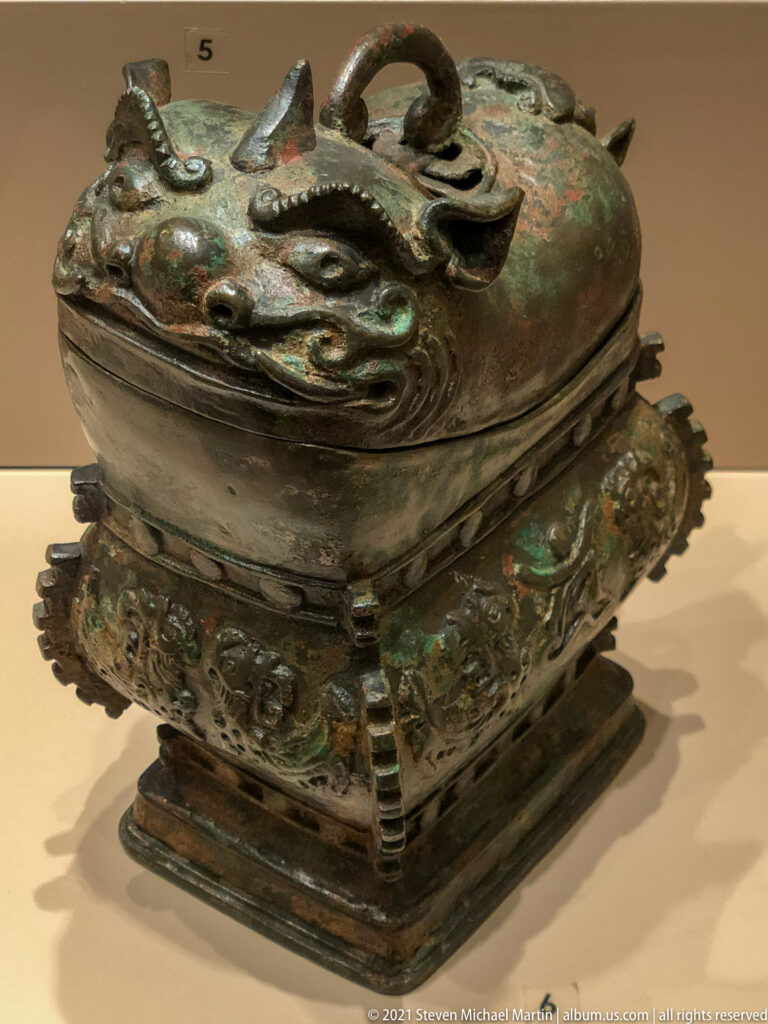 Square bronze covered vessel with modeled animal forms on the lid, relief decoration, and patches of green corrosion.