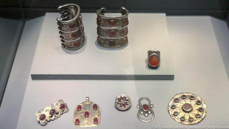 Group of silver-colored cylindrical ornaments, pendants, and circular fittings set with oval red stones in a display case.